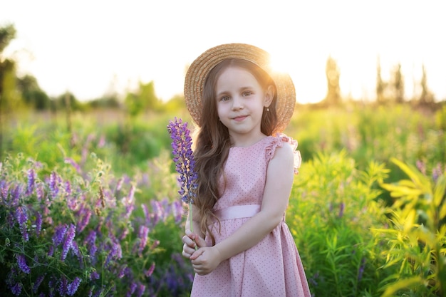 Smiling little girl in straw hat holds lupine flower in her hands at a field