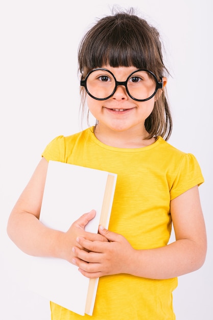 Smiling little girl wearing yellow t-shirt and round black glasses holding a book in her hands on white background