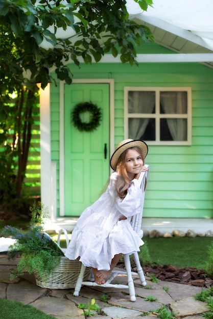 Smiling little girl in white dress and hat sitting with basket
wildflowers on green lawn in garden