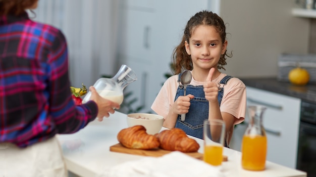 Smiling little girl with curly hair shows the thumbs having healthy breakfast together with mother in the kitchen.