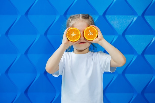 Smiling little girl with pigtails in a white T-shirt on a blue background with a cut orange in her hands. Children's emotions, fun