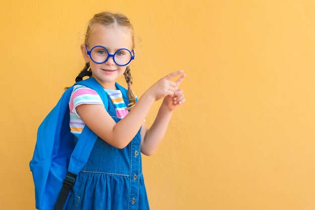 眼鏡の学校制服のバックパックを着た笑顔の小さな学校の子供の女の子は、コピースペースで指を指して注目を集めています。学校の広告学用品や文房具に戻る