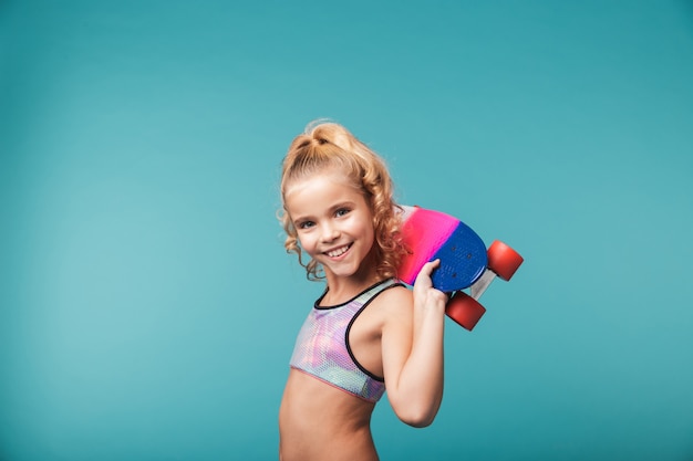 Улыбающаяся маленькая спортивная девочка играет со скейтбордом, изолированным на синей стене