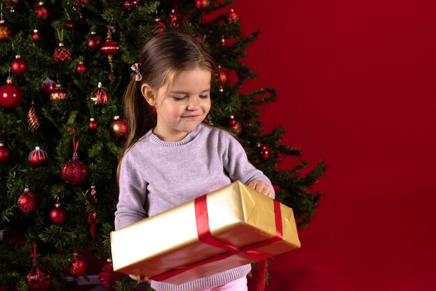 Smiling little toddler girl with gift box next to Christmas tree