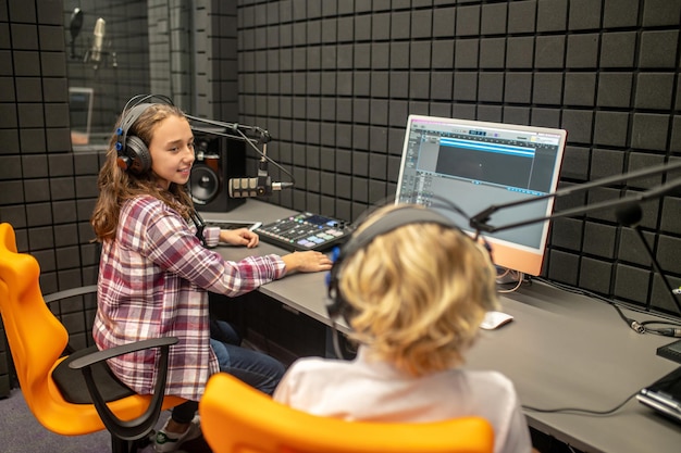 Foto sorridente bella ragazza dai capelli lunghi in una camicia a quadri seduta alla scrivania accanto a un ragazzo biondo in studio