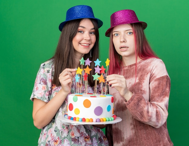 Sorridente guardando la telecamera ragazze che indossano il cappello da festa con in mano una torta