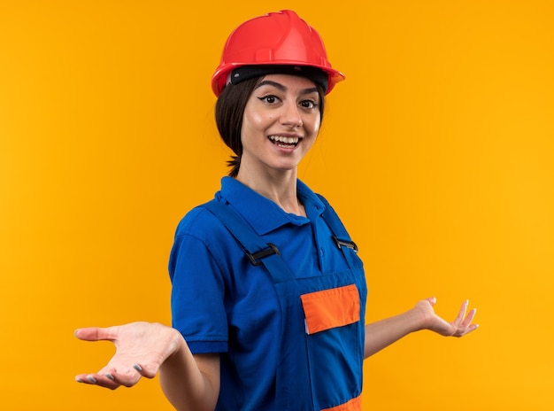 Sorridente guardando la telecamera giovane donna costruttore in uniforme allargando le mani