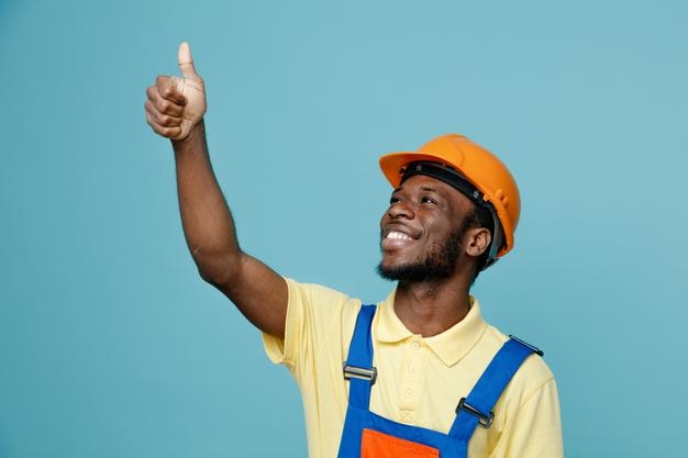 Smiling looking at side showing thumbs up young african american builder in uniform isolated on blue background