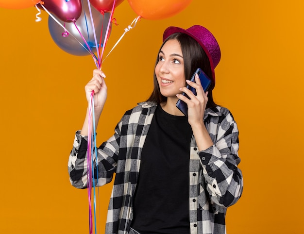 Улыбаясь, глядя в сторону, молодая красивая женщина в партийной шляпе с воздушными шарами говорит по телефону, изолированному на оранжевой стене
