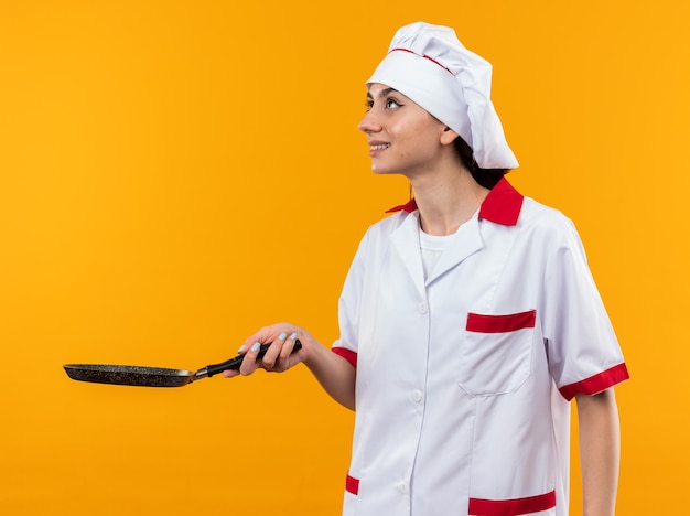 Sorridente guardando la giovane bella donna in uniforme da chef tenendo fuori la padella sul lato isolata sulla parete arancione