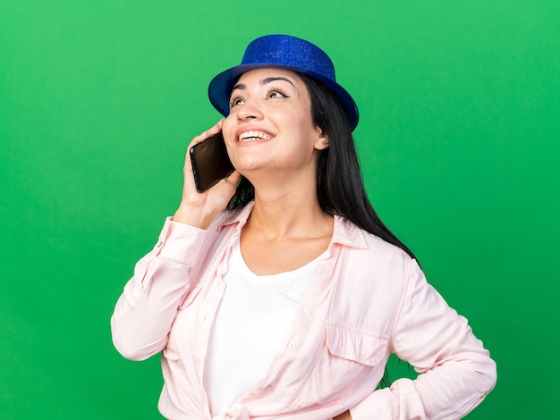 Smiling looking up young beautiful woman wearing party hat speaks on phone putting hand on hip isolated on green wall