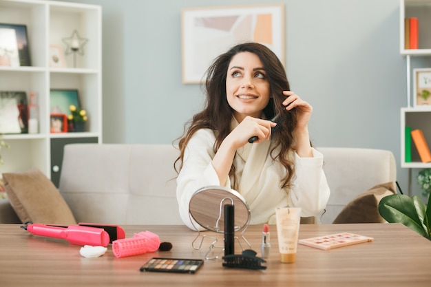 Smiling looking up young girl combing hair sitting at table with makeup tools in living room