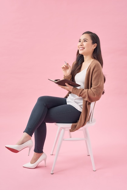 Smiling lovely girl writing in a diary while sitting on a chair isolated over pink wall