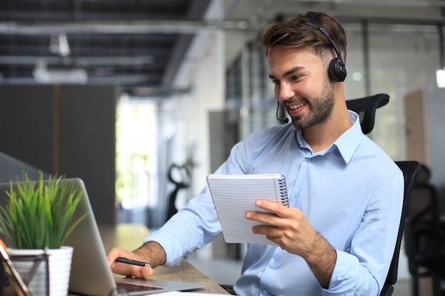 Sorridente consulente aziendale maschio con le cuffie seduto in un ufficio moderno, videochiamata guardando lo schermo del laptop. assistenza clienti dell'agente del servizio clienti dell'uomo che parla di chat online.