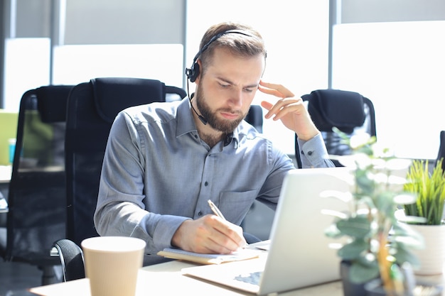 Sorridente operatore di call center maschio con cuffie seduto in un ufficio moderno, consultando informazioni online in un laptop, cercando informazioni in un file per essere di aiuto al cliente