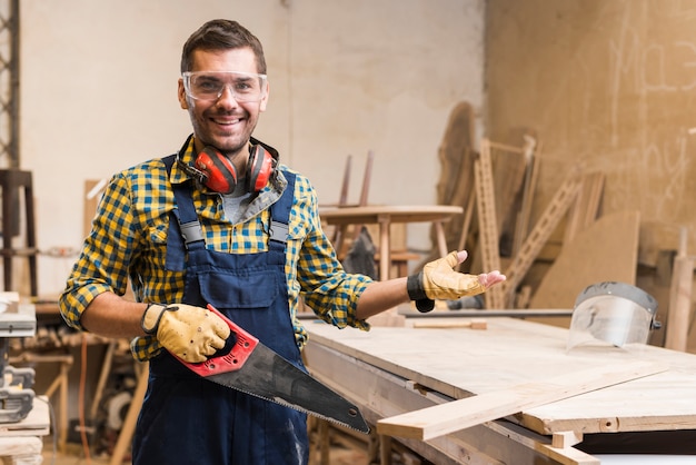 Foto vetri di sicurezza d'uso sorridenti del carpentiere maschio che tengono gesturing della sega a mano