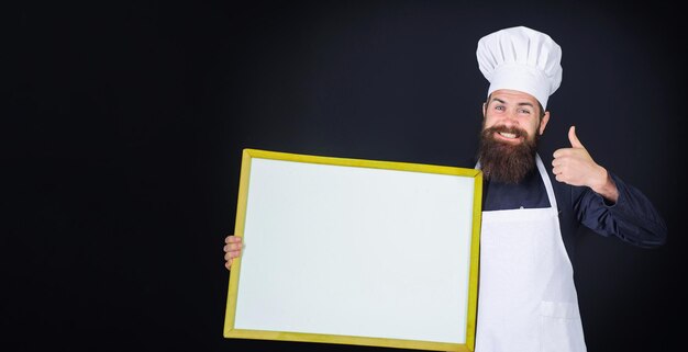 빈 메뉴와 함께 제복을 입은 요리사를 엄지손가락으로 보여주는 빈 보드와 함께 웃고 있는 남성 요리사 요리사 또는 베이커