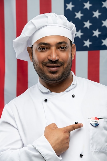 Smiling male chef of restaurant in uniform pointing at vote insignia on chest while standing against stars-and-stripes