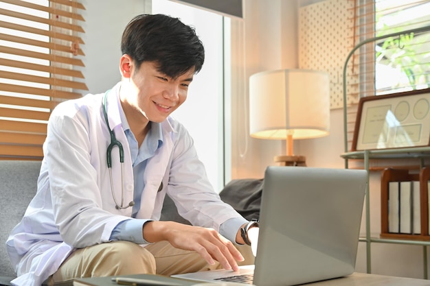 웃고 있는 남성 의사가 환자와 멀리 떨어져 의사소통을 하거나 노트북으로 가정 건강 치료를 위한 온라인 상담을 제공합니다.