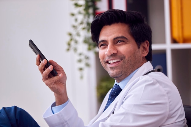 Smiling Male Doctor Or GP Wearing White Coat Sitting At Desk In Office With Mobile Phone