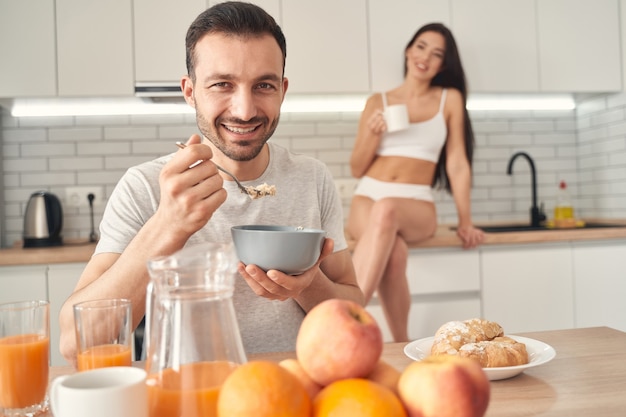 Smiling male eating oat flakes on breakfast with pleasure. Pretty wife holding cup of tea in hand