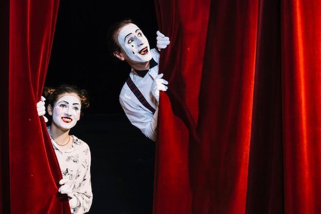 Smiling male and female mime artist peeking from red curtain