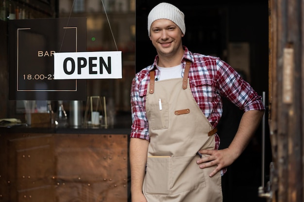 バーの笑顔の男性オーナーが屋外に立っています。彼はオープンに来るようにみんなを招待しています。