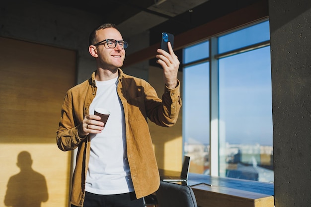 Smiling male worker in casual clothes looking away while talking on mobile phone and drinking coffee during break in modern creative workspace