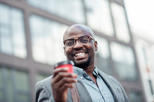 Uomo sorridente. impiegato afroamericano barbuto con gli occhiali che sorride e beve caffè