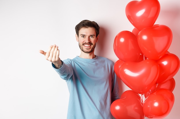 Smiling man beckon you to come closer, follow me gesture, taunting his lover move forward, have romantic surprise, standing near red balloon on valentines day, white background