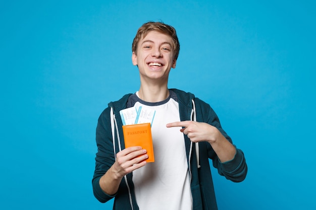 Smiling man in casual clothes holding, pointing index finger on passport and boarding pass ticket isolated on blue wall. People sincere emotions, lifestyle concept. 