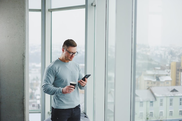 Smiling man in casual clothes standing near window with coffee\
and looking at smartphone in modern workspace with big window\
during daytime