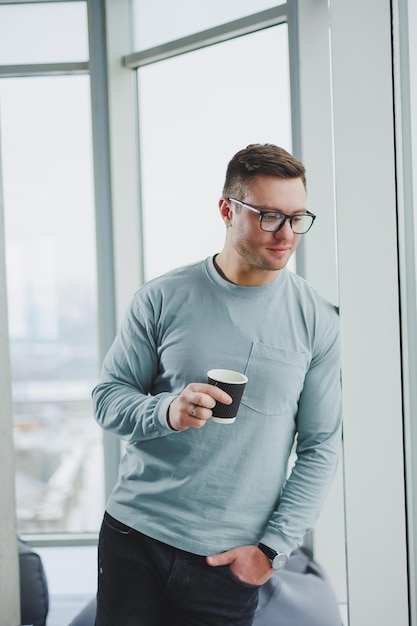 Smiling man in casual clothes standing near window with coffee in modern work area with big window during daytime