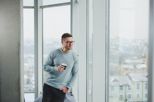 Smiling man in casual clothes standing near window with coffee\
in modern work area with big window during daytime