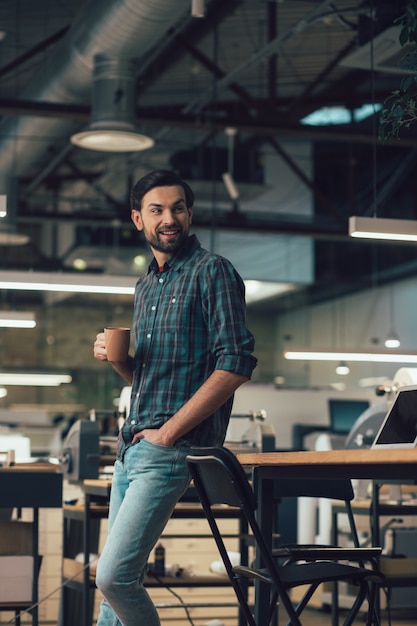 Smiling man in casual clothes standing next to the table with a mug in his hand