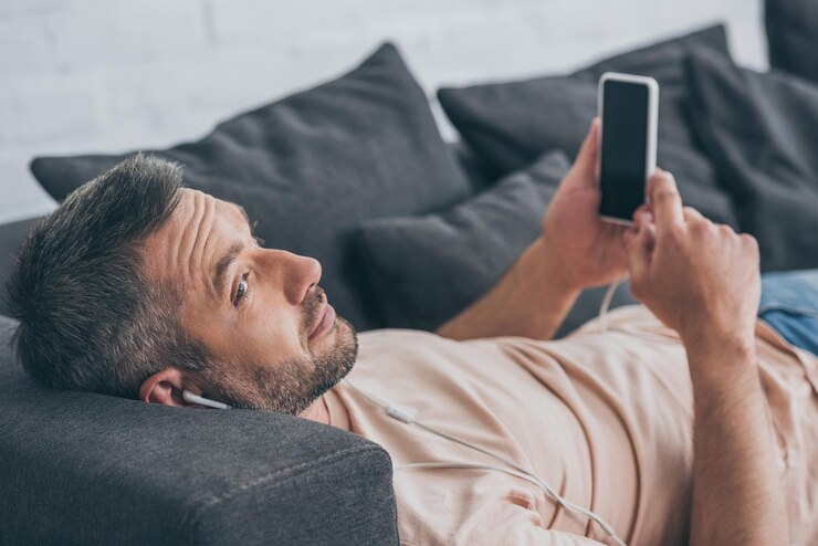 Photo smiling man in earphones lying on sofa and holding smartphone with blank screen