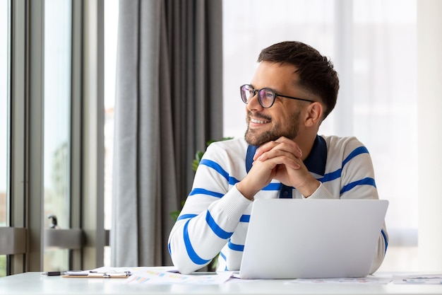 L'uomo sorridente con gli occhiali si siede alla scrivania in ufficio, navigando in internet wireless sul dispositivo portatile, il lavoratore maschio felice si rilassa durante la pausa di lavoro