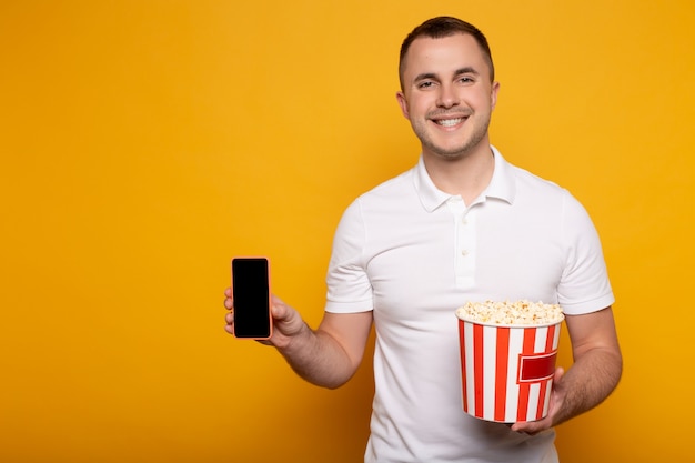 Smiling man holding bucket with popcorn and smartphone on yellow