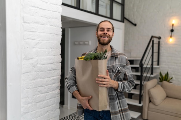 Улыбающийся мужчина с бумажной сумкой для покупки продуктов, полной свежих здоровых овощей в руке, стоящий дома