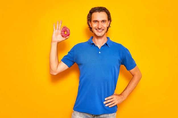 Smiling man holds donut in his hand in form of an OK sign he is dressed in casual clothes blue polo