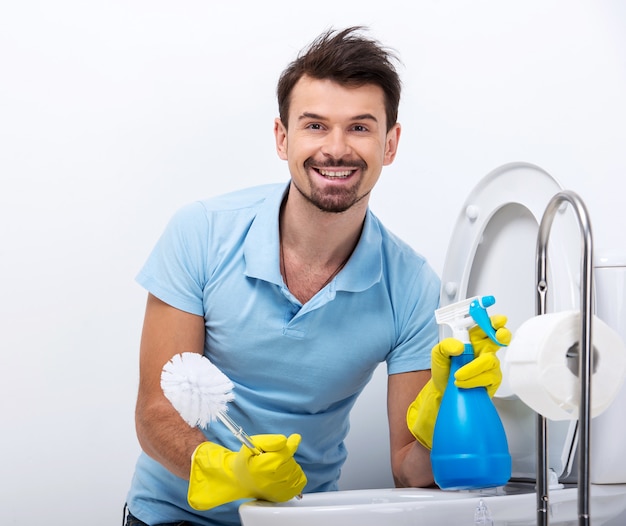 L'uomo sorridente sta pulendo la toilette con il detergente e la spazzola dello spruzzo.