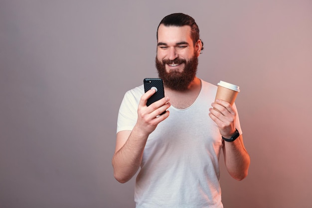 Smiling man is surfing the internet on phone while holding a coffee cup