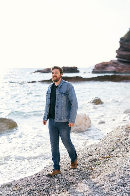 Smiling man in jeans and a denim jacket walks along the pebble beach near the water looking into the