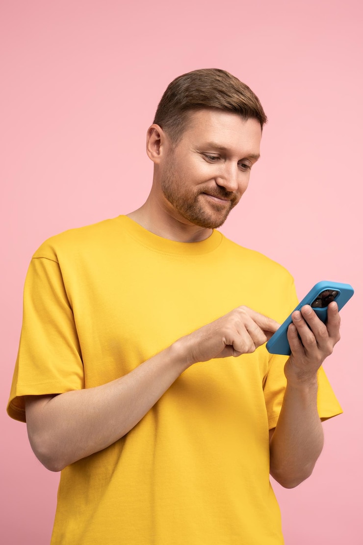 Photo smiling man looking at smartphone screen browsing in internet chatting with friends over pink wall