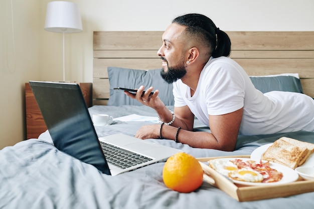 Smiling man lying on bed with breakfast tray, working on laptop and recording voice message for colleague