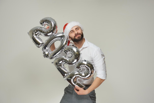 A smiling man in a red velvet Santa hat is hugging silver balloons in the shape of 2022 gently. A happy guy with a beard at a New year party.