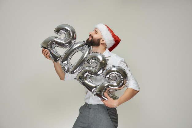 A smiling man in a red velvet santa hat is hugging silver\
balloons in the shape of 2022 gently. a happy guy with a beard at a\
new year party.