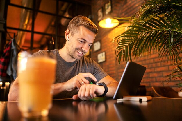 Photo smiling man sitting at cafe and typing on smart watch while expecting video call on tablet