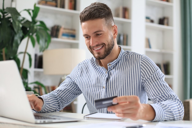 Smiling man sitting in office and pays by credit card with his laptop.