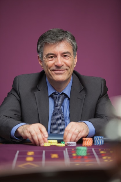 Smiling man sitting at table in a casino while playing roulette and grabbing chips